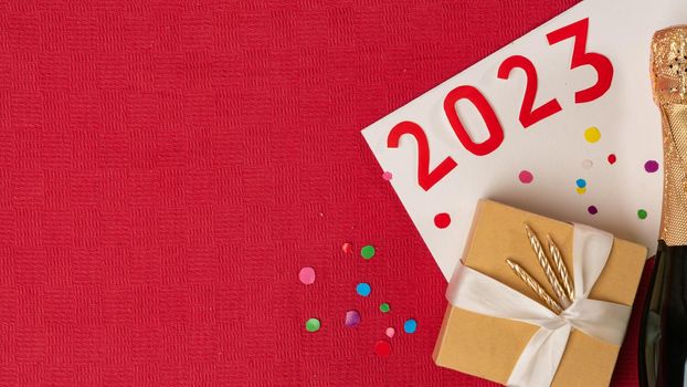 New Year 2023 with a bottle of champagne and a gift box on a red background with confetti. High quality photo