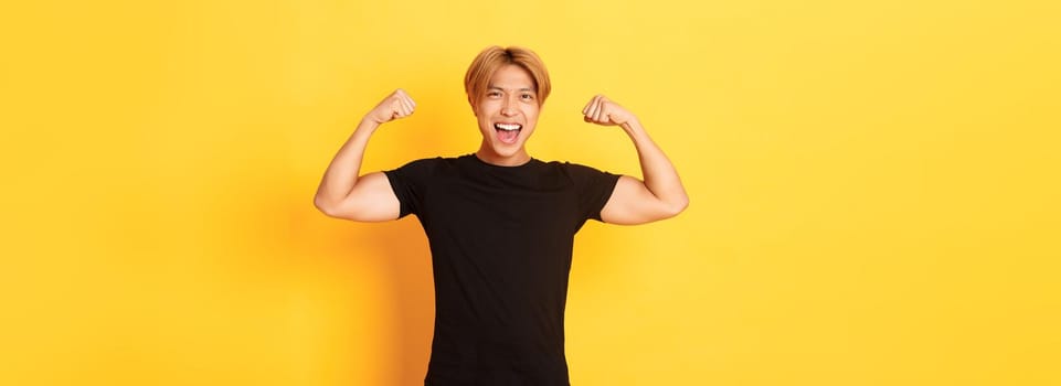 Portrait of happy and confident asian blond guy, flexing biceps, showing strong big muscles, standing yellow background.