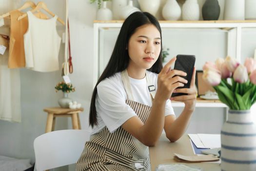 Asian female, small stock business owner holding phone product photography checking commercial shipping delivery order on smartphone using mobile app technology.