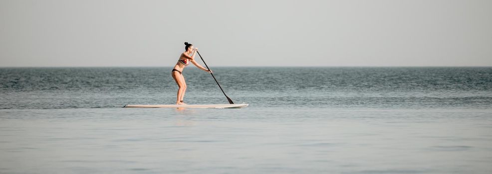 Silhouette of woman standing, surfing on SUP board, confident paddling through water surface. Idyllic sunset or sunrise. Sports active lifestyle at sea or river.