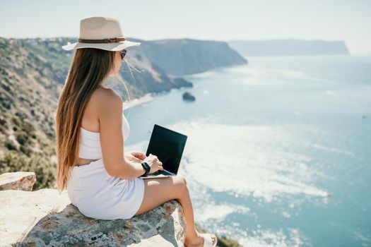 Successful business woman in yellow hat working on laptop by the sea. Pretty lady typing on computer at summer day outdoors. Freelance, travel and holidays concept.