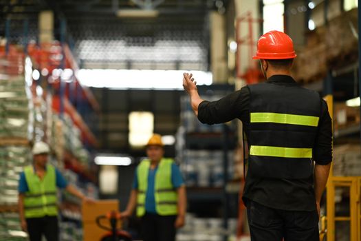 Male manager wearing hardhat and reflective jacket checking inventory production stock control in warehouse.