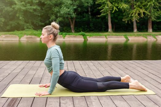 A beautiful woman lies in a gray top and leggings does yoga in summer on a wooden platform by a pond in the park. Copy space. Side view
