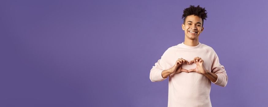 Portrait of young happy, romantic lovely boyfriend express his love and sympathy to dearest person, show heart sign and beaming smile, like this girl and trying to confess feelings, purple background.