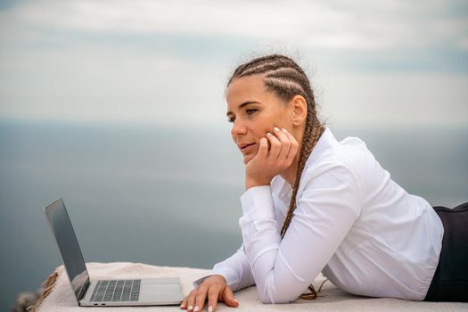 A woman is lying and typing on a laptop keyboard on a terrace with a beautiful sea view. Wearing a white blouse and black skirt. Freelance travel and vacation concept, digital nomad