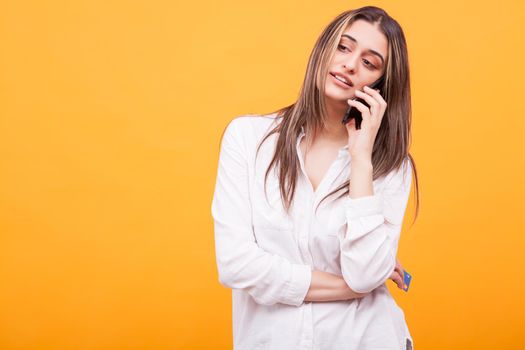Bored girl in white shirt talking on the phone and holding credit card over yellow background. Casual dressed