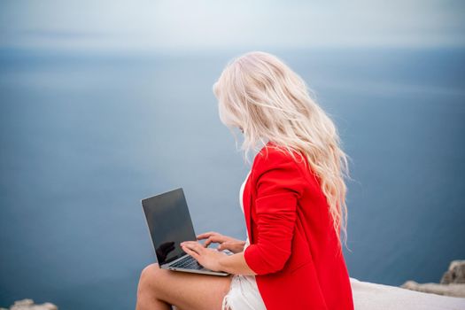 A woman is typing on a laptop keyboard on a terrace with a beautiful sea view. Freelancing, digital nomad, travel and vacation concept