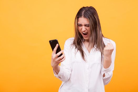 Portrait of a happy young girl holding mobile phone and celebrating isolated over yellow background. Showing satisfaction