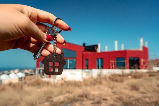 A woman's hand holds a house key against the backdrop of a house under construction. Real estate agent. Buying a house, apartment