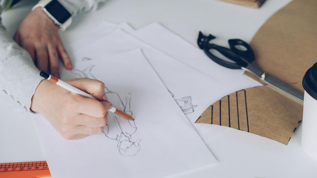 Close-up of fashion designer is drawing lines on clothing sketch. Woman is busy and concentrated on work.