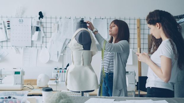 Young woman is measuring mannequin parts with tape-measure while her female colleague is entering this information in smartphone. Productive cooperation in tailoring business concept.