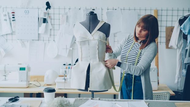 Young seamstress is measuring clothing pinned to tailoring dummy with measure-tape. Attractive woman is concentrated and thoughtful. Studio is light, modern, with many sewing tools and items.