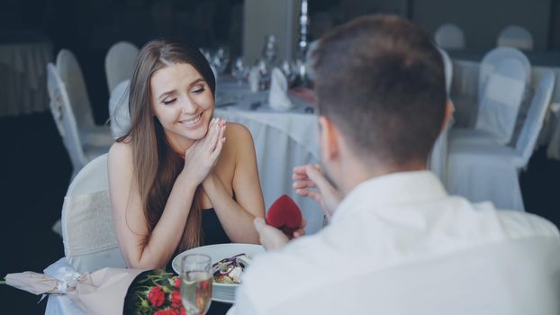 Handsome young guy is proposing to surprised pretty woman in beautiful dress, then putting engagement ring on her finger and kissing her hand during romantic date in restaurant.