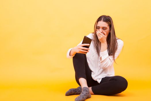 Girl looking shocked at her phone while sitting down over yellow background. Facial expression