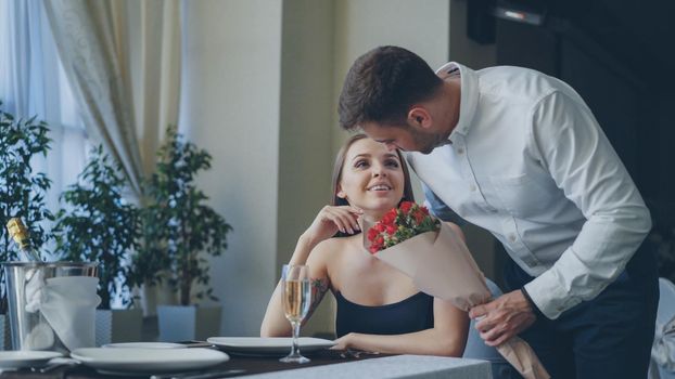 Beautiful woman is waiting for her boyfriend in nice restaurant, he is coming and giving her flowers, kissing her. Happy girl is smelling flowers and smiling with pleasure.