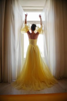 A woman's silhouette in a golden luxurious dress against the background of a window holds a curtain with her hands. Elegant lady in a yellow long silk dress with bare back, back view