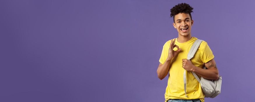 Back to school, university concept. Portrait of young handsome hipster guy, student with backpack assure test be good, show okay sign, chill and relax no worried, all ok, purple background.