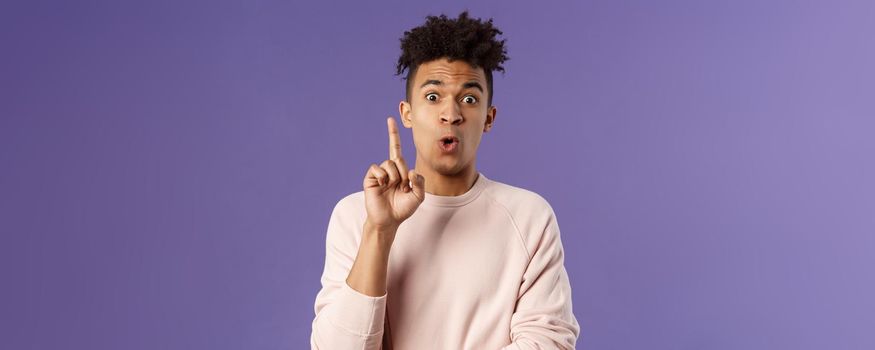 Got it, have plan. Portrait of excited happy young man, rejoicing as being striked with great idea, raise index finger in eureka gesture, figured out something, have suggestion, purple background.