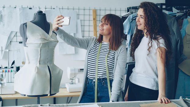 Two creative female designers are making selfie with smart phone while standing beside tailoring dummy in modern workplace. Attractive women are smiling and posing.