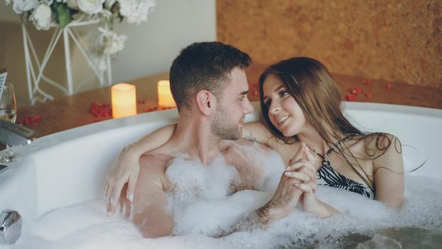 Loving young people in bathing suits are talking, hugging and touching hands in hot bathtub in spa salon. Romantic relationship, passion and wellness concept.