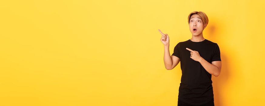 Portrait of impressed asian guy looking and pointing fingers upper left corner, standing yellow background.