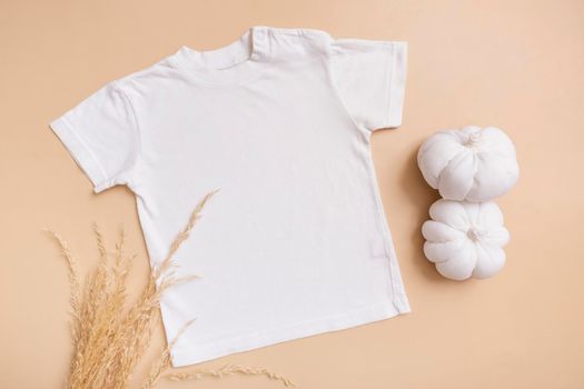 White baby t-shirt top view. Mock-up for logo, text or design on beige background. Flat lay child clothes with decorative pumpkins.