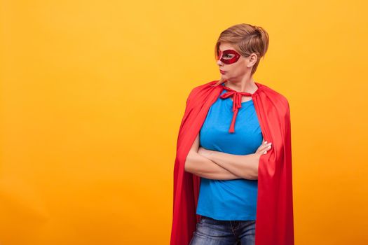 Woman dressed like superhores standing proudly and looking away over yellow background. Woman super powers. Red cape. red mask