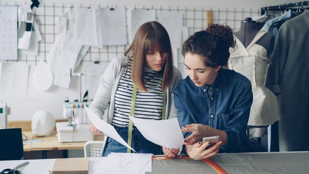 Creative female fashion designers are busy watching sketches working near sewing desk. Young women are talking and checking smartphone. Modern tailor's shop in background.