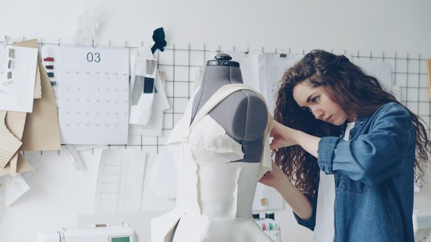 Young pretty seamstress is adjusting clothing on tailoring dummy with sewing pins. Fashionable women's garments, sketches on wall, tailoring tools are visible. Everyday work at tailor shop concept.