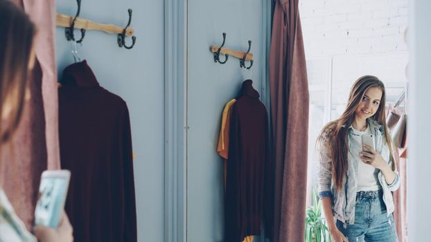 Pretty young girl is making mirror selfie with smartphone while standing in fitting room in clothes store. She is posing, turning and touching her long brown hair with lovely smile.