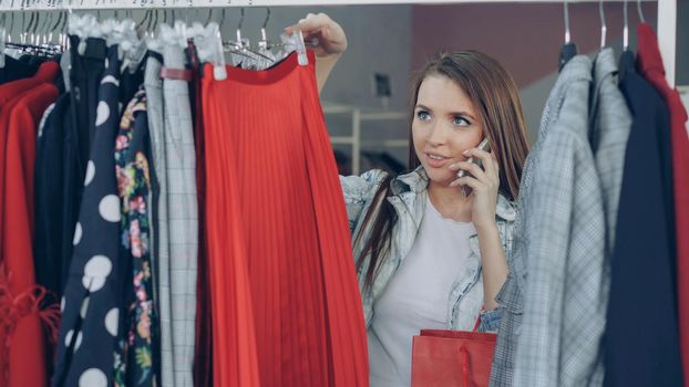 Attractive blond girl choosing fine clothes in modern shop and talking cheefully on mobile phone. Close-up shot of colourful skirts and jackets on hangers.