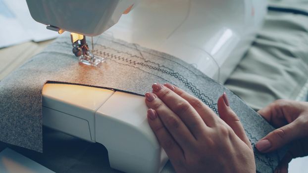Close-up shot of working modern sewing machine, fabric and manicured female hand. Clothes manufacturing process concept. Light soft colours, beautiful stitches.