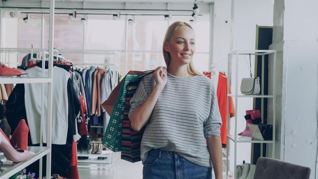 Dolly shot of blond girl walking between shelves and rails in large store. She is carrying lots of bags, smiling carelessly and looking at fine modern clothes around her.