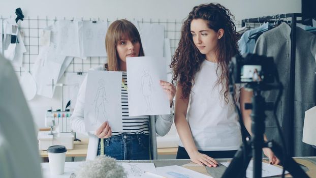 Creative bloggers tailors recording video about new sketches. Geirls are greeting followers, showing drawings and talking emotionally. Camera, garments and tailoring items are visible.