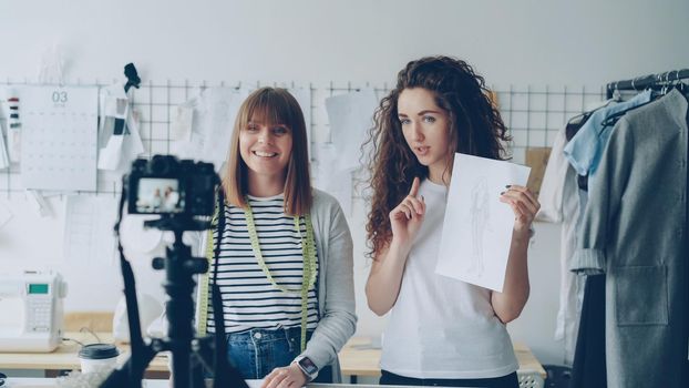 Young attractive vloggers clothing designers are recording video for their vlog. Women are showing garment sketches, gesturing and talking emotionally in tailoring studio.