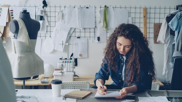 Young woman fashion designer is drawing ladies' garment sketch at desk in modern workshop. Mannequin, sewing items, to-go coffee are visible. Creativity in clothing manufacturing concept.