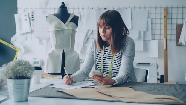 Creative clothing designer is looking at smartphone screen and drawing sketch while working in modern tailor shop at sewing table. Woman is busy and involved in process.