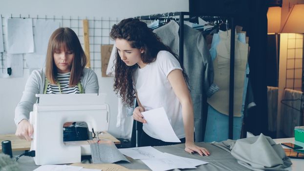 Attractive female dressmakers are looking at sketches and working with sewing machine, then checking stitches and adjusting equipment. Professional teamwork concept.