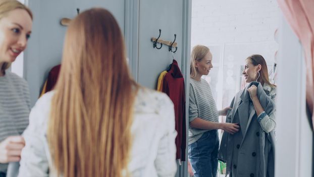 Pretty young woman is checking fashionable coat in fitting room with her friend helping her to appraise garment. They are talking, gesturing and looking at clothing in large mirror.