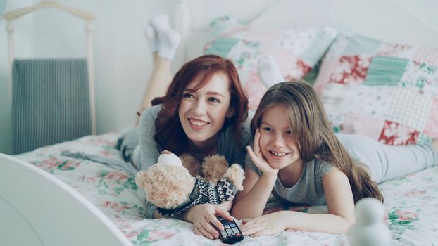 Laughing happy mother and cute daughter watching funny cartoon movie on TV while lying on bed at home in morning have fun at cozy bedroom