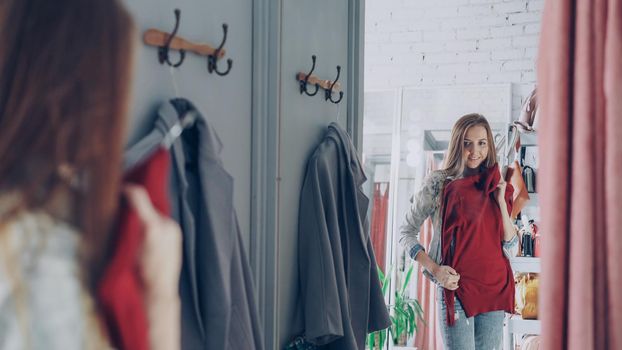 Mirror shot of young lady choosing clothes in fitting room. Girl is trying jumper checking size and length while standing opposite large mirror. Nice clothing store in background.