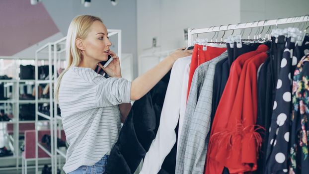 Female customer is slowly going through fashionable clothes on hanger in spacious shop. Other customers are moving around with lots of bright clothes and shoes in the background.