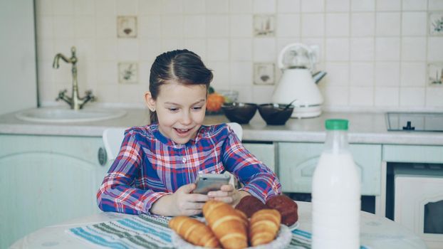 Little cute girl playing smartphone at morning while sitting at table in kitchen. Childhood, people, and technology concept