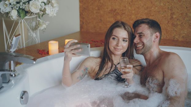 Adorable young couple is taking selfie with sparkling champagne glasses using smartphone while bathing in bathtub. They are smiling, kissing and posing looking at camera.