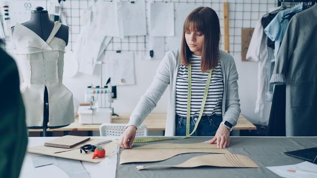 Young pretty seamstress is measuring clothing patterns with tape-measure at studio table while working in her modern tailor's shop. Getting ready for sewing clothes concept.