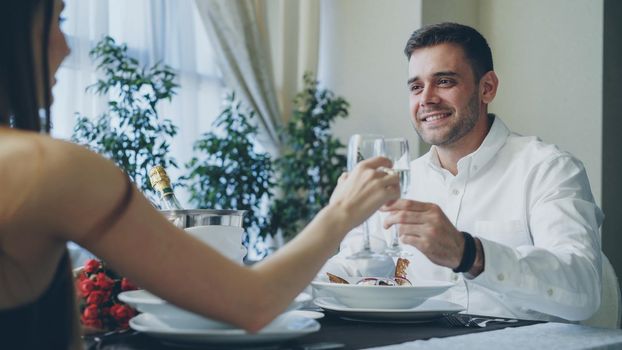 Young handsome man is talking to his girlfriend clinking glasses and drinking champagne while having dinner in classy restaurant.