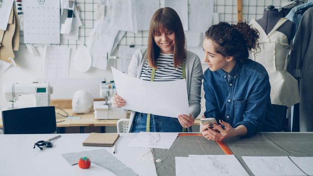 Attractive young women working in design industry are comparing sketches and talking about them while working in modern tailor shop. Productive teamwork concept.