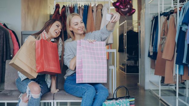 Cute girls are sitting and making selfie with colourful paper bags using smartphone in luxurious women's clothing boutique, then watching pictures together. They are smiling and laughing carelessly.