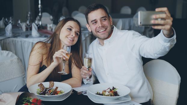 Attractive loving couple is taking selfie with champagne glasses using smartphone while having romantic dinner in restaurant. They are smiling and posing looking at camera.