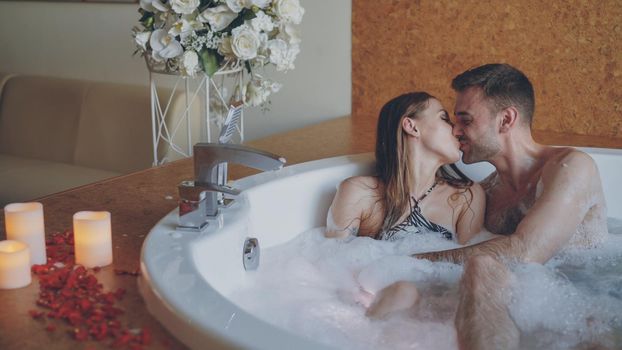 Attractive cheerful couple in hot tub is playing blowing foam, kissing and enjoying romantic moments. Rose petals, beautiful flowers and burning candles are visible.
